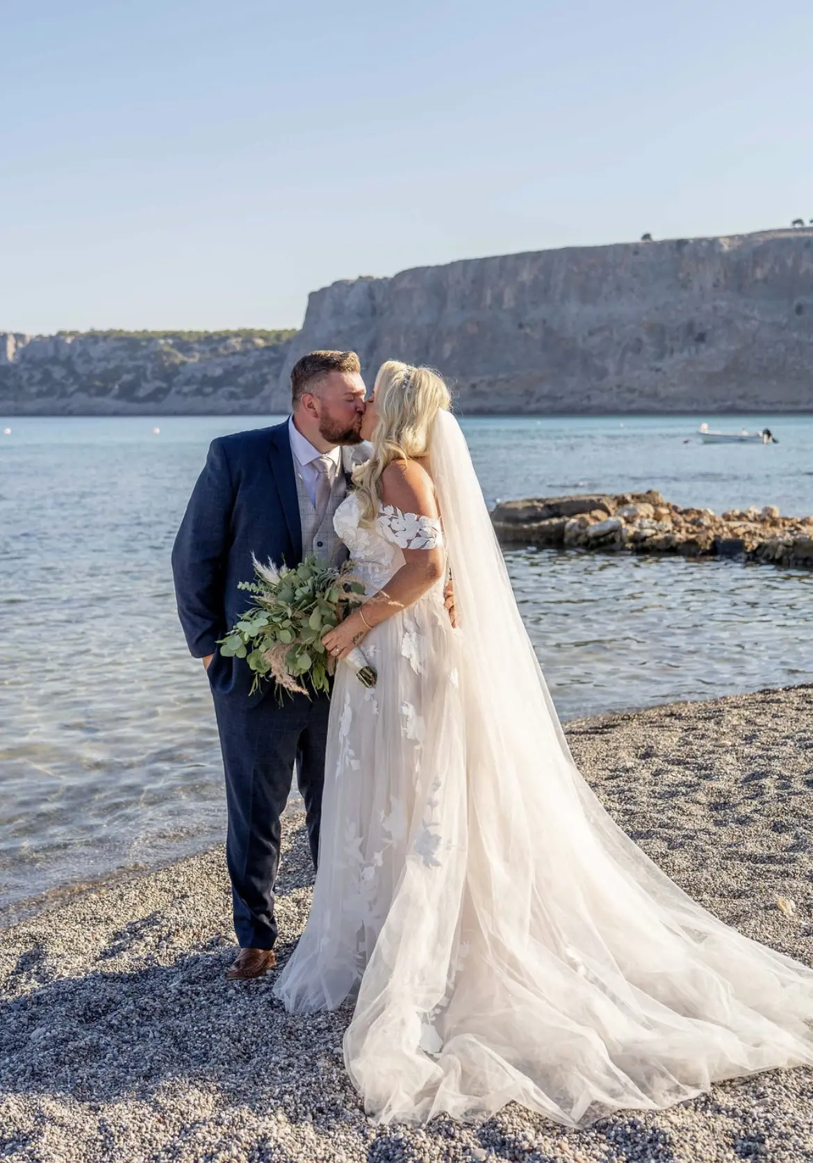 Groom Kissing Bride by Water in Greece Wedding Wearing Boho Wedding Dress Called Hattie Lane Marie by Rebecca Ingram.