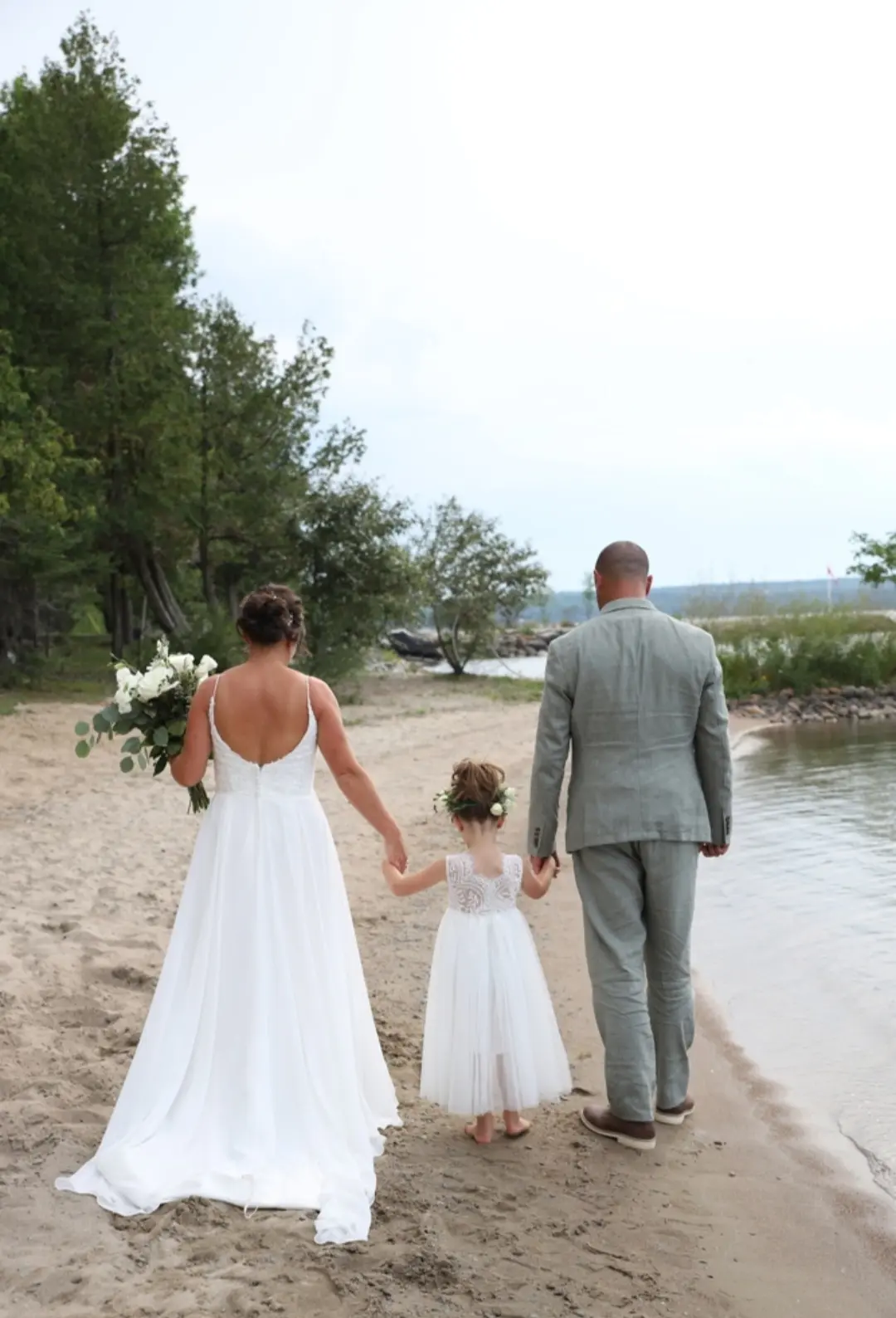 Bride Wearing Simple Chiffon Wedding Dress Called Marta Lynette By Rebecca Ingram Walking Away With Flower Girl And Groom Near Water