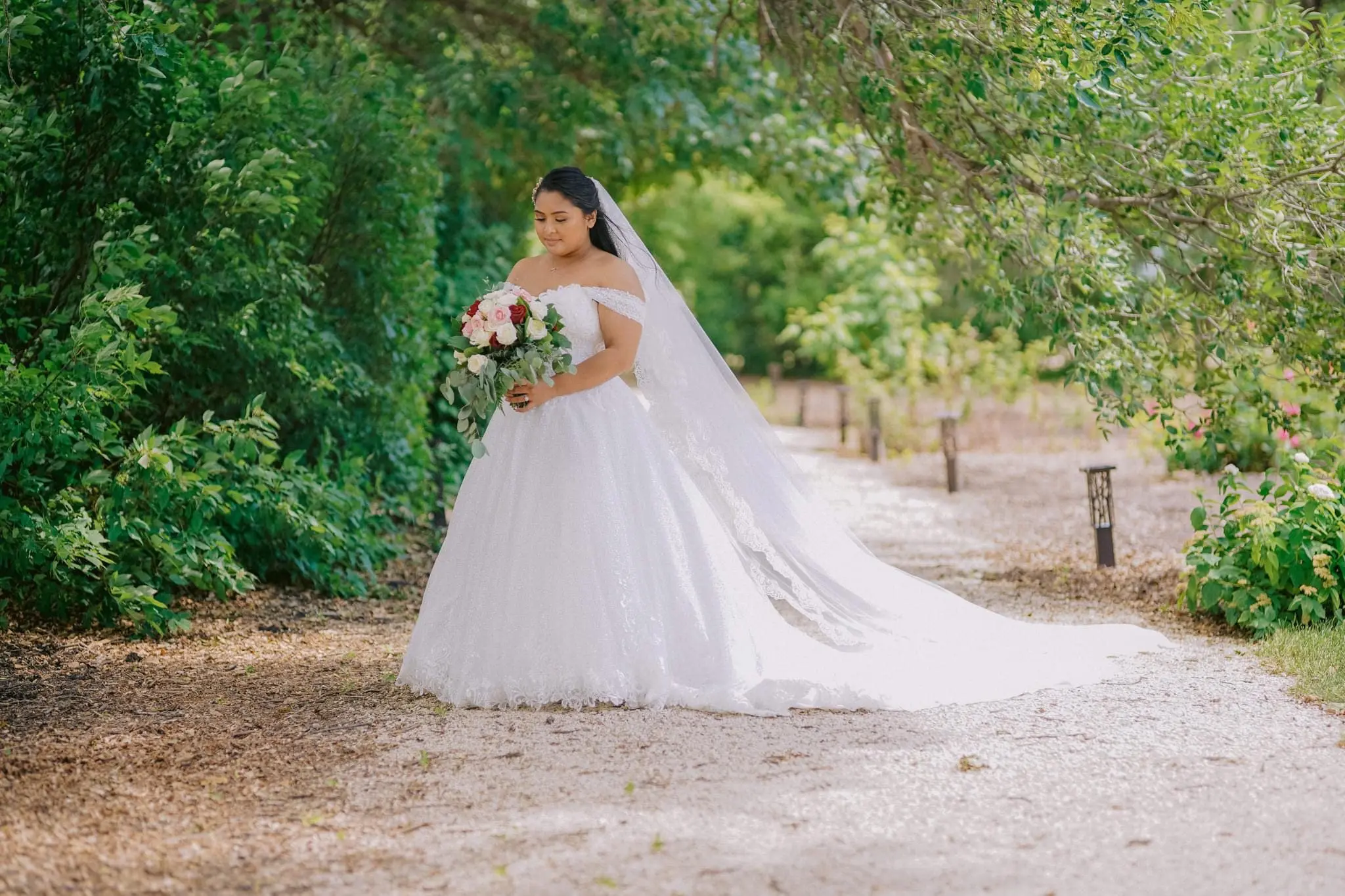Bride stood in trees looking at her bouquet of mixed pink and ivory hues wearing wedding dress called Tristyn.
