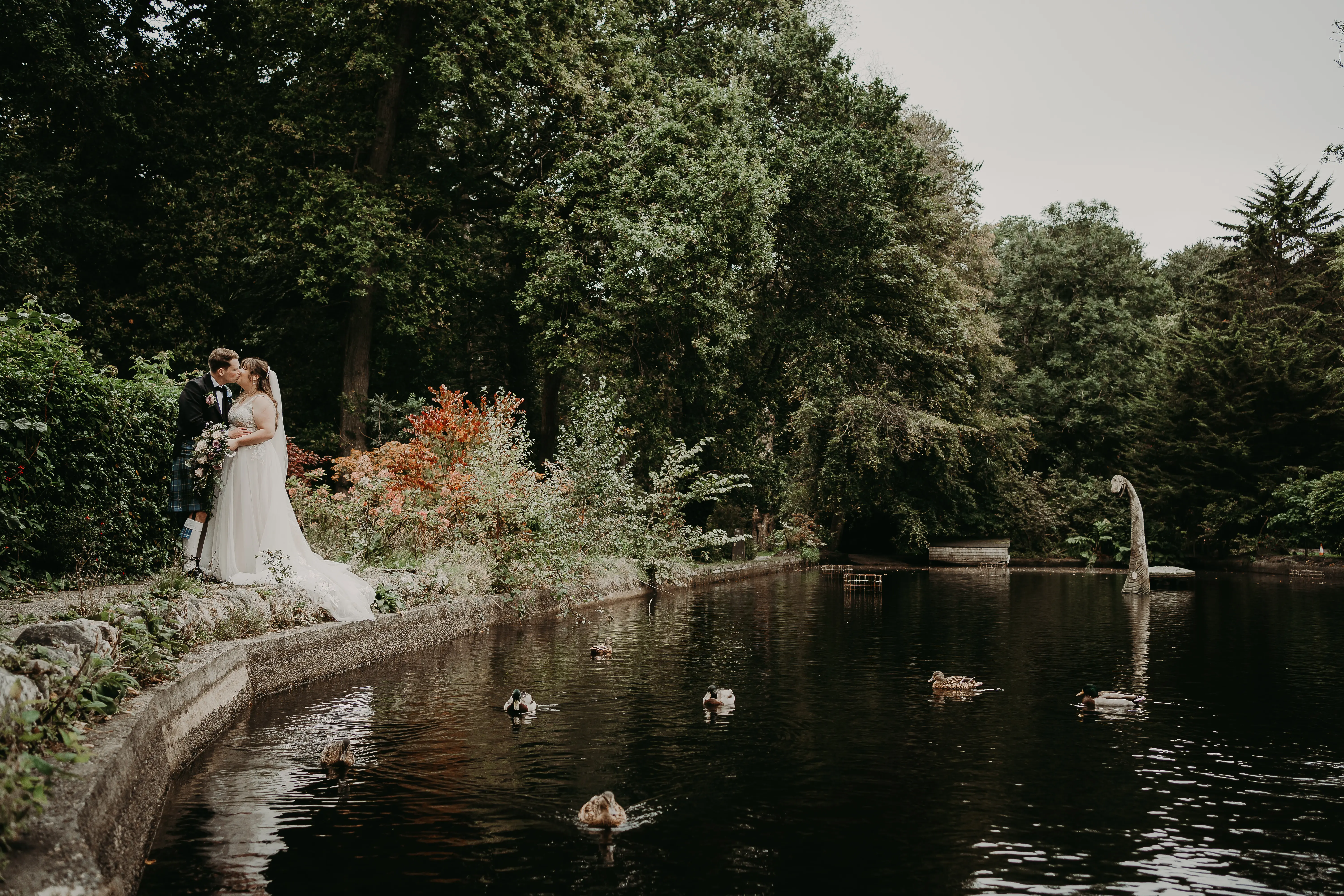 Newlyweds kissing near a lake with ducks and faux loch ness monster.