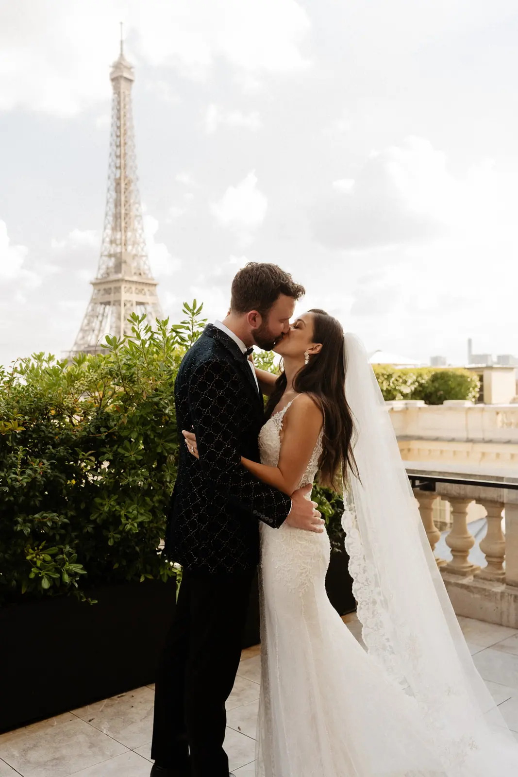 Groom Kissing Bride In Front Of Eiffel Tower Wearing a Beaded Lace Sheath Bridal Gown Called Barrett By Sottero And Midgley