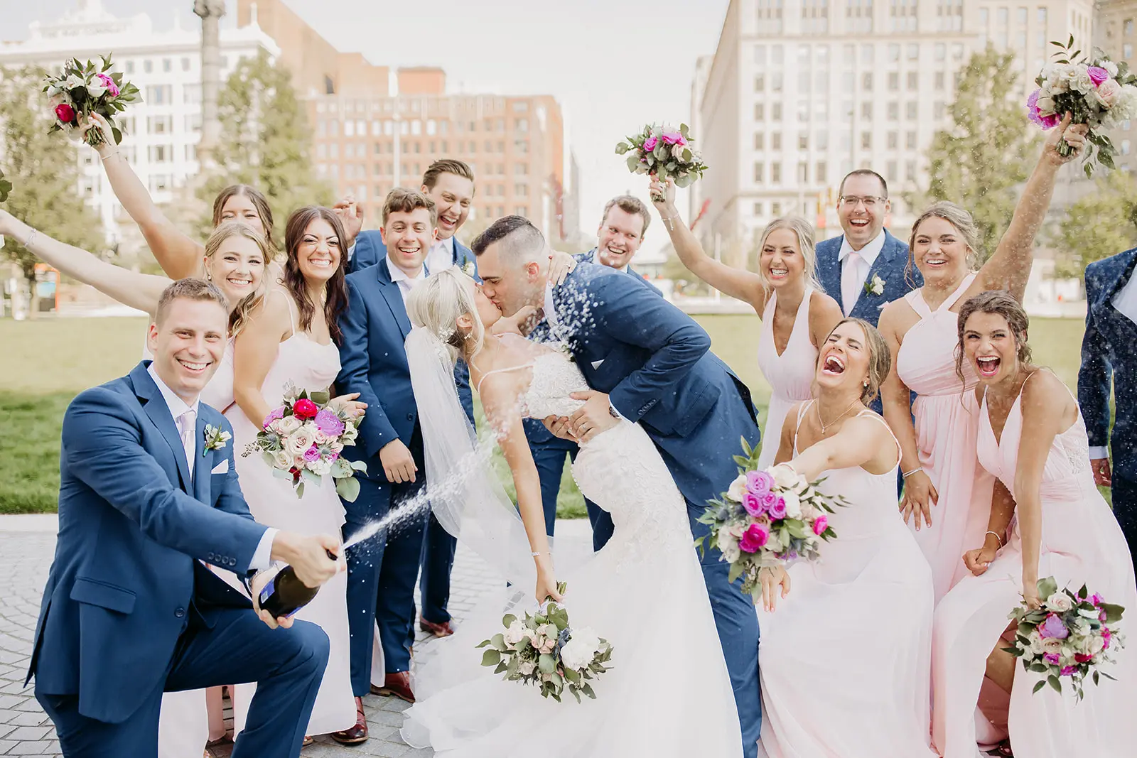 Groom kissing bride as wedding party celebrates with spraying champagne and cheering.