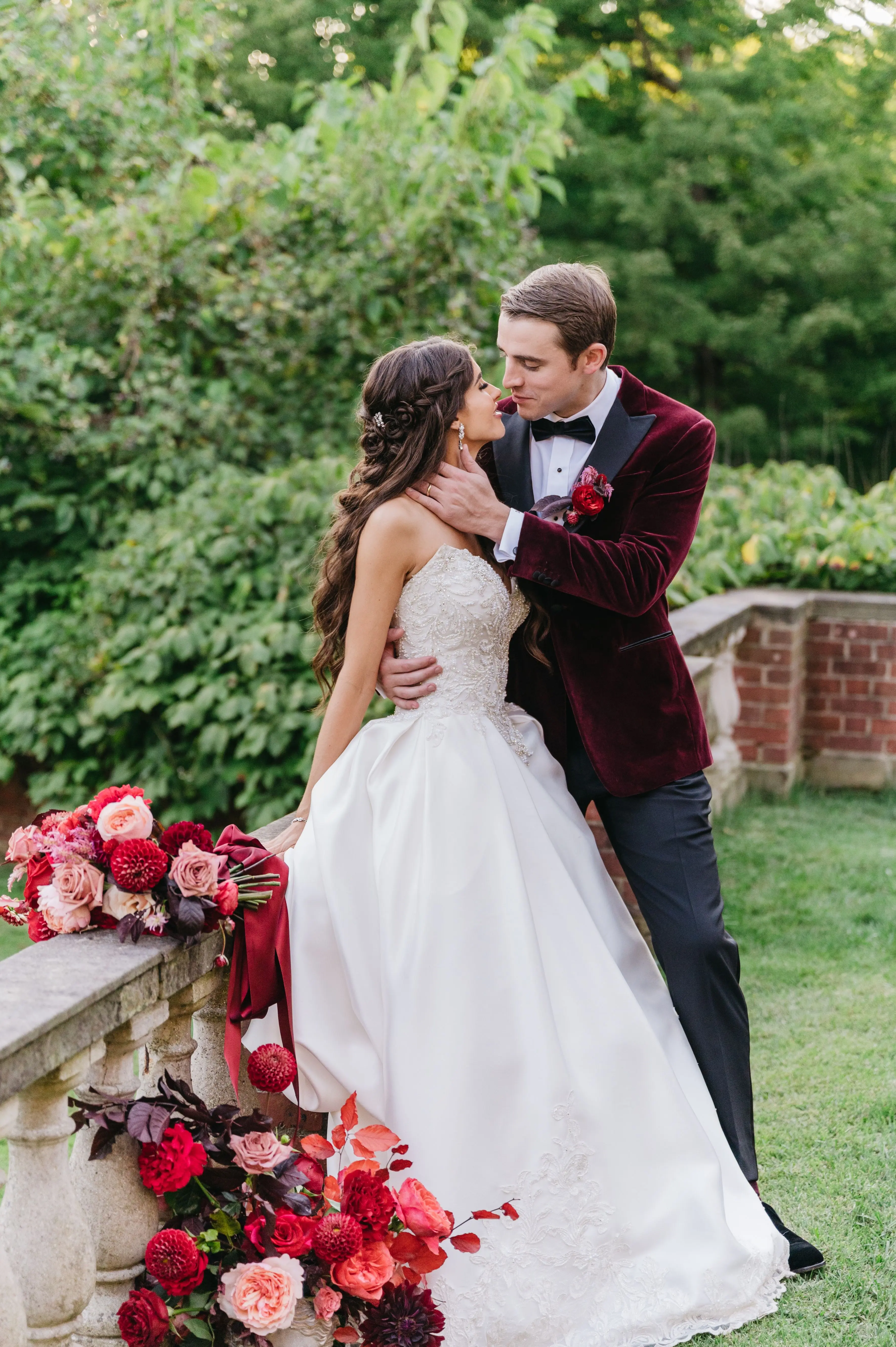 Bride In Satin Ballgown Wedding Dress Called Kimora By Sottero And Midgley With Red And Pink Florals And Groom In Red Velvet Suit