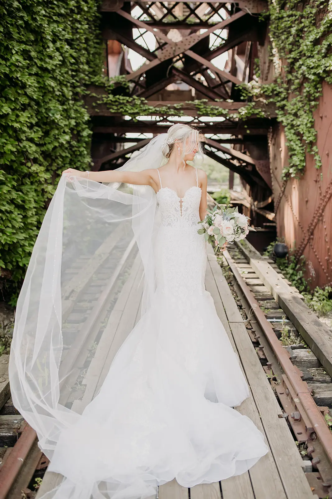 Bride stood on railway track holding out her veil wearing wedding dress called Alistaire.
