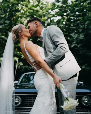 Bride wearing Fontaine by Maggie Sottero kissing her husband in front of a car