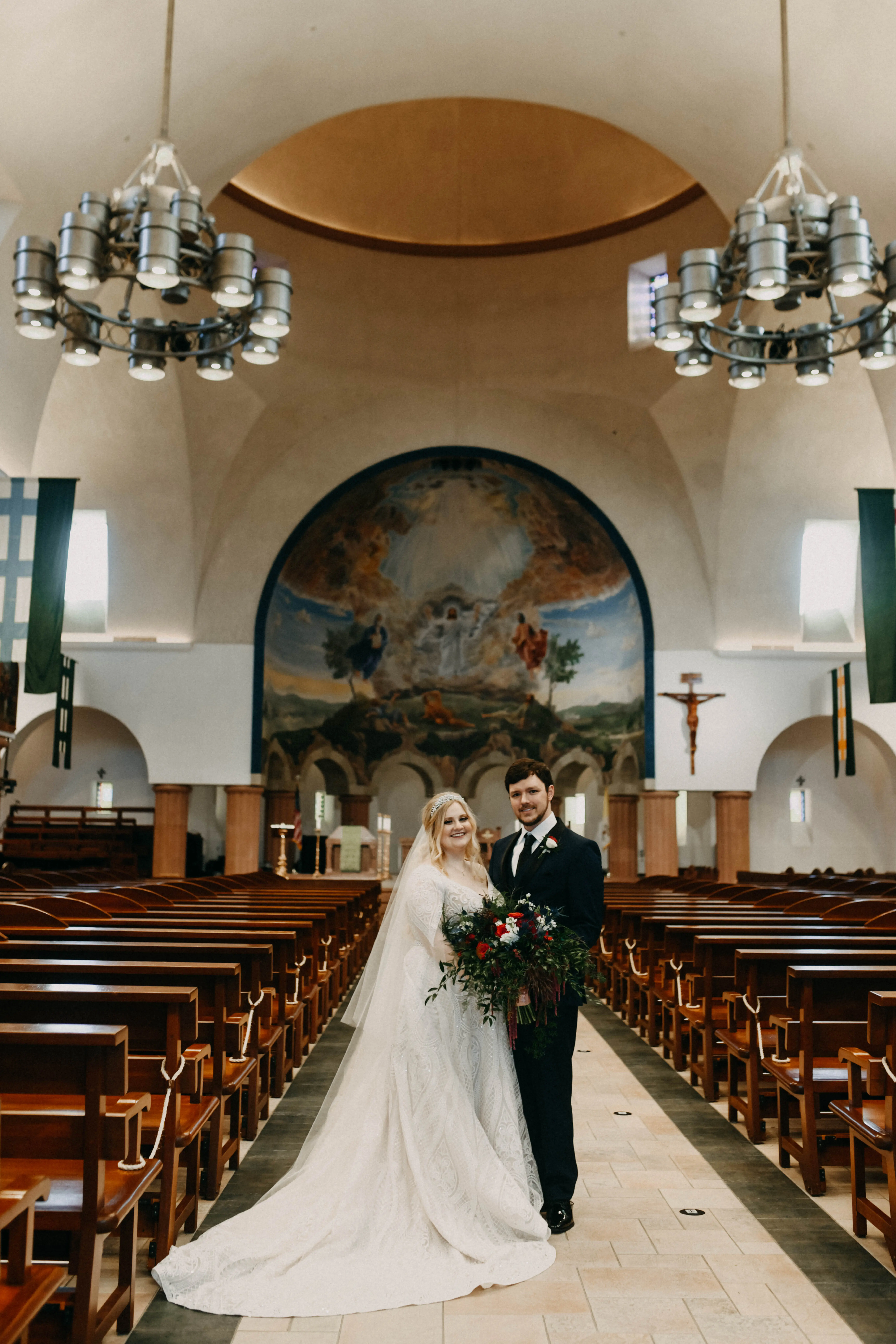 Bride and groom stood in the aisle of picturesque church bride holding large red and green bouquet.