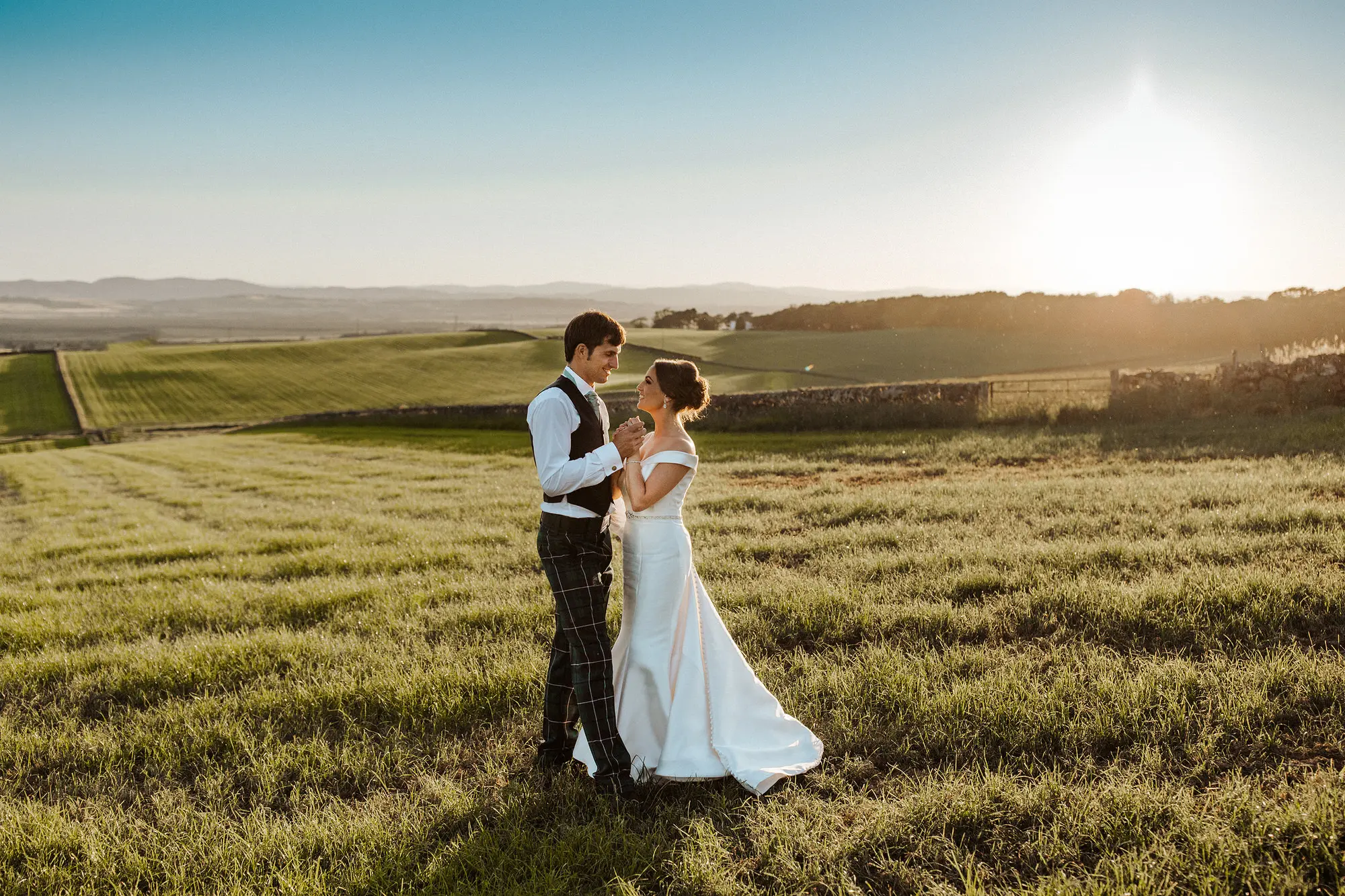 Newlyweds holding hands facing each other on a beautiful field at sunset.