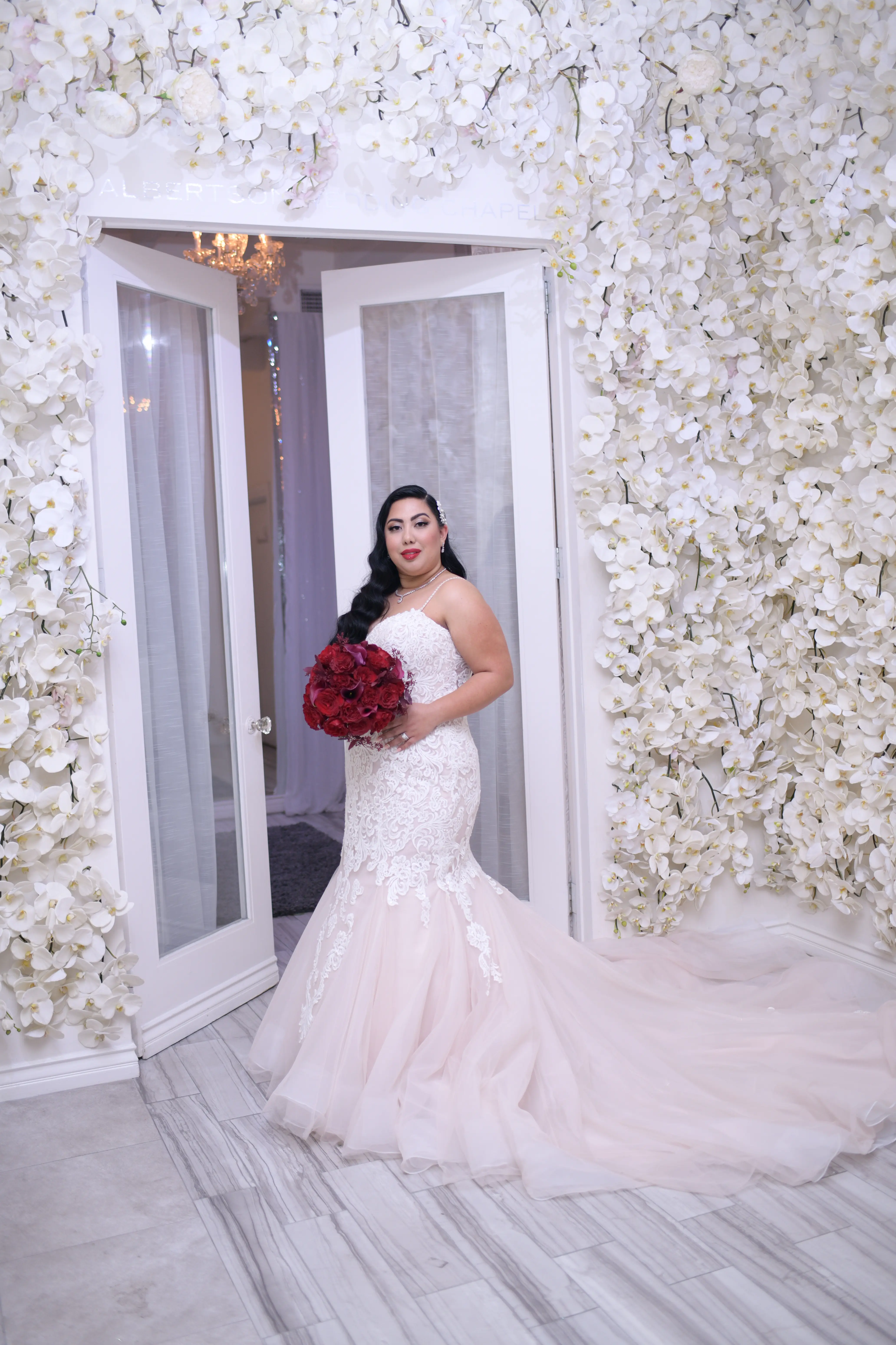 Bride stood infront of doorway with the walls covered in ivory flowers and bride wearing wedding dress called Alistaire Lynette.