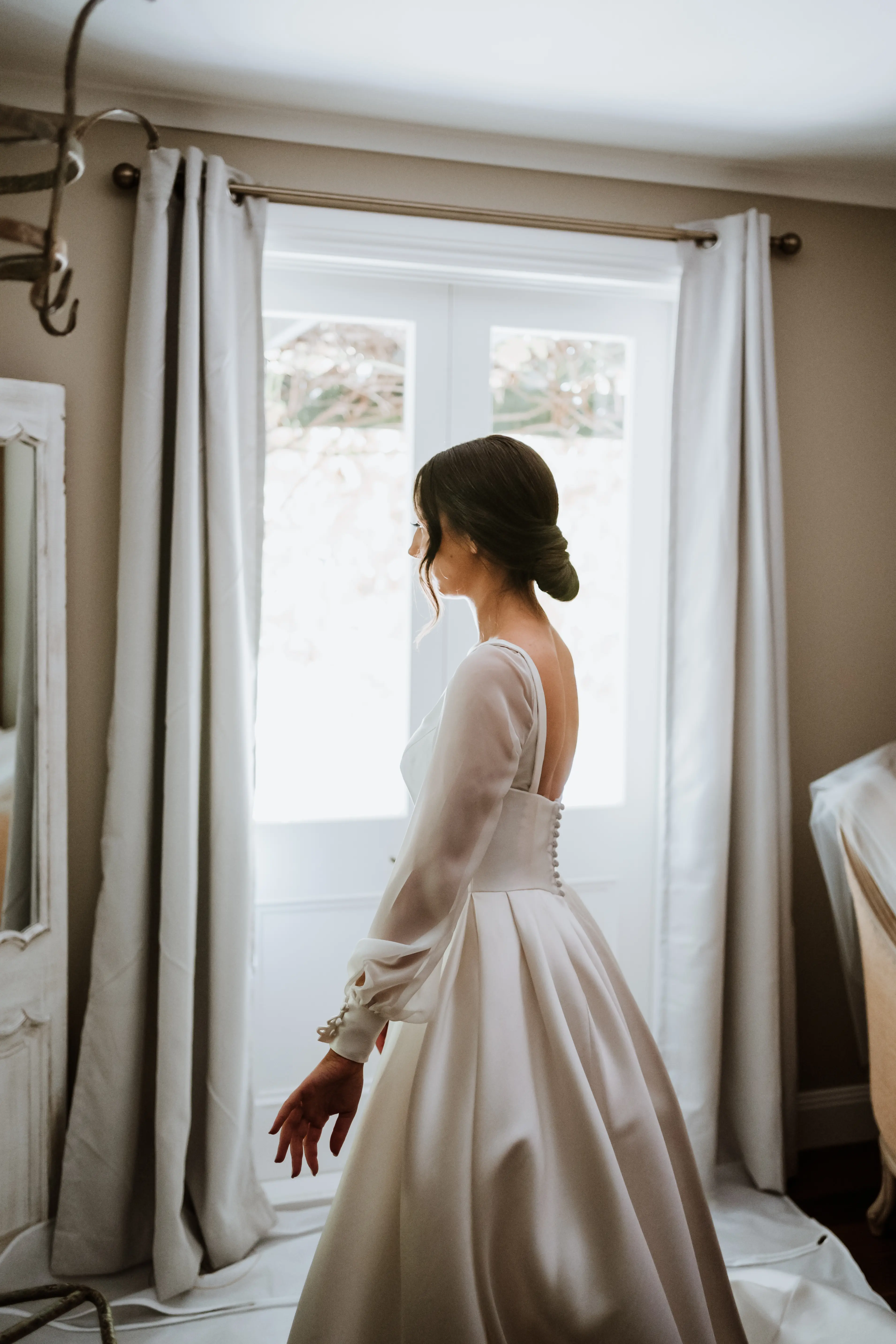 Bride wearing wedding dress called Selena with personalized sleeves stood by open window.