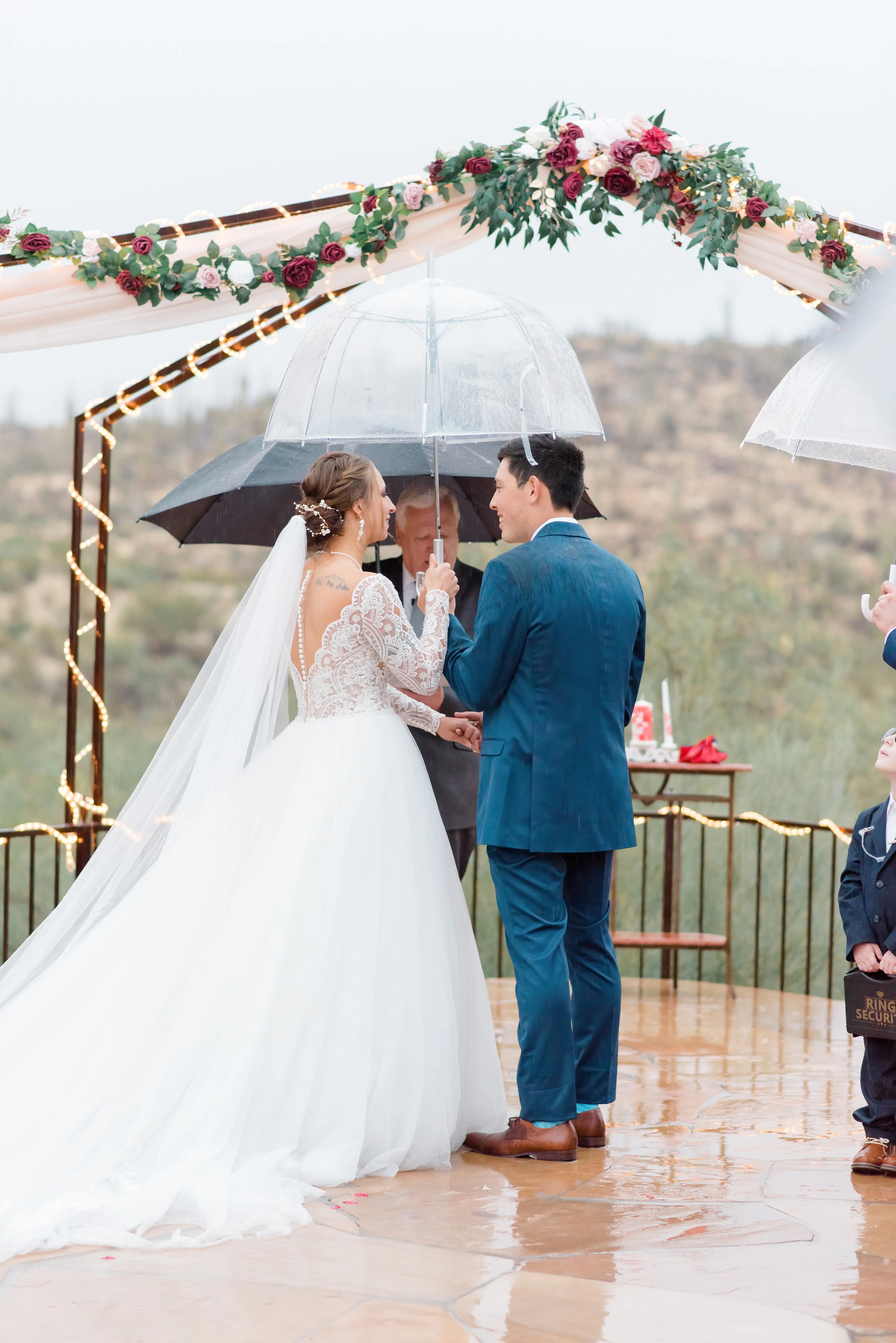 Groom Under Umbrella with with Bride on Rainy Wedding Day. Bride in Romantic Long Sleeve Ball Gown Wedding Dress with Lace Motifs on Nude Bodice and Tulle Skirt Called Mallory Dawn by Maggie Sottero