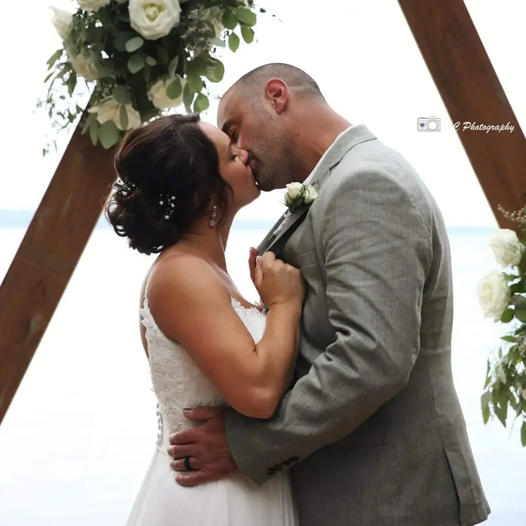 Bride Wearing Simple Chiffon Wedding Dress Called Marta Lynette By Rebecca Ingram Kissing Groom Under Triangle Arch With Flowers