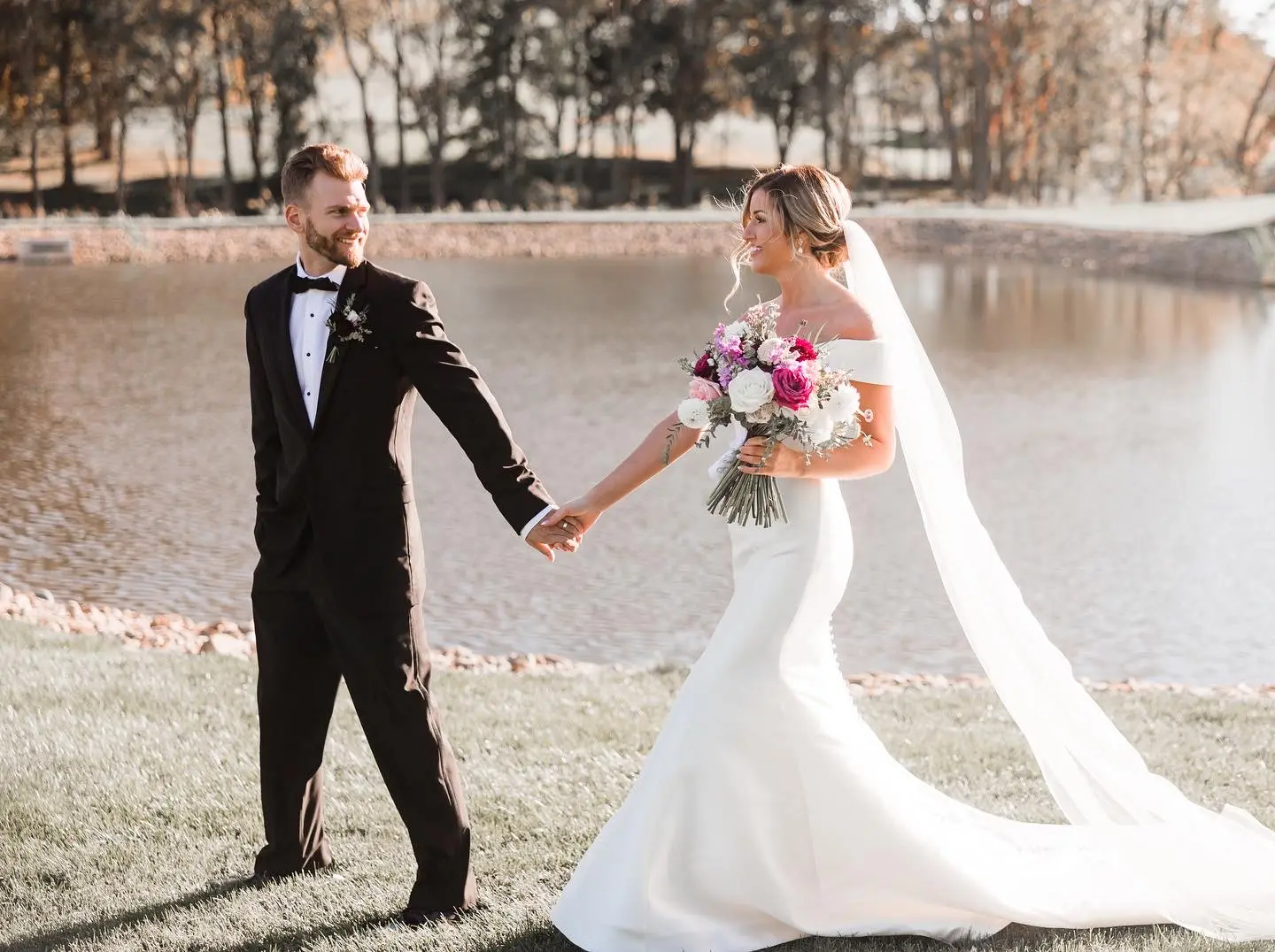 Groom Walking With Bride In Off The Shoulder Fit And Flare Wedding Dress Named Josie By Rebecca Ingram Holding Pink Flower Bouquet