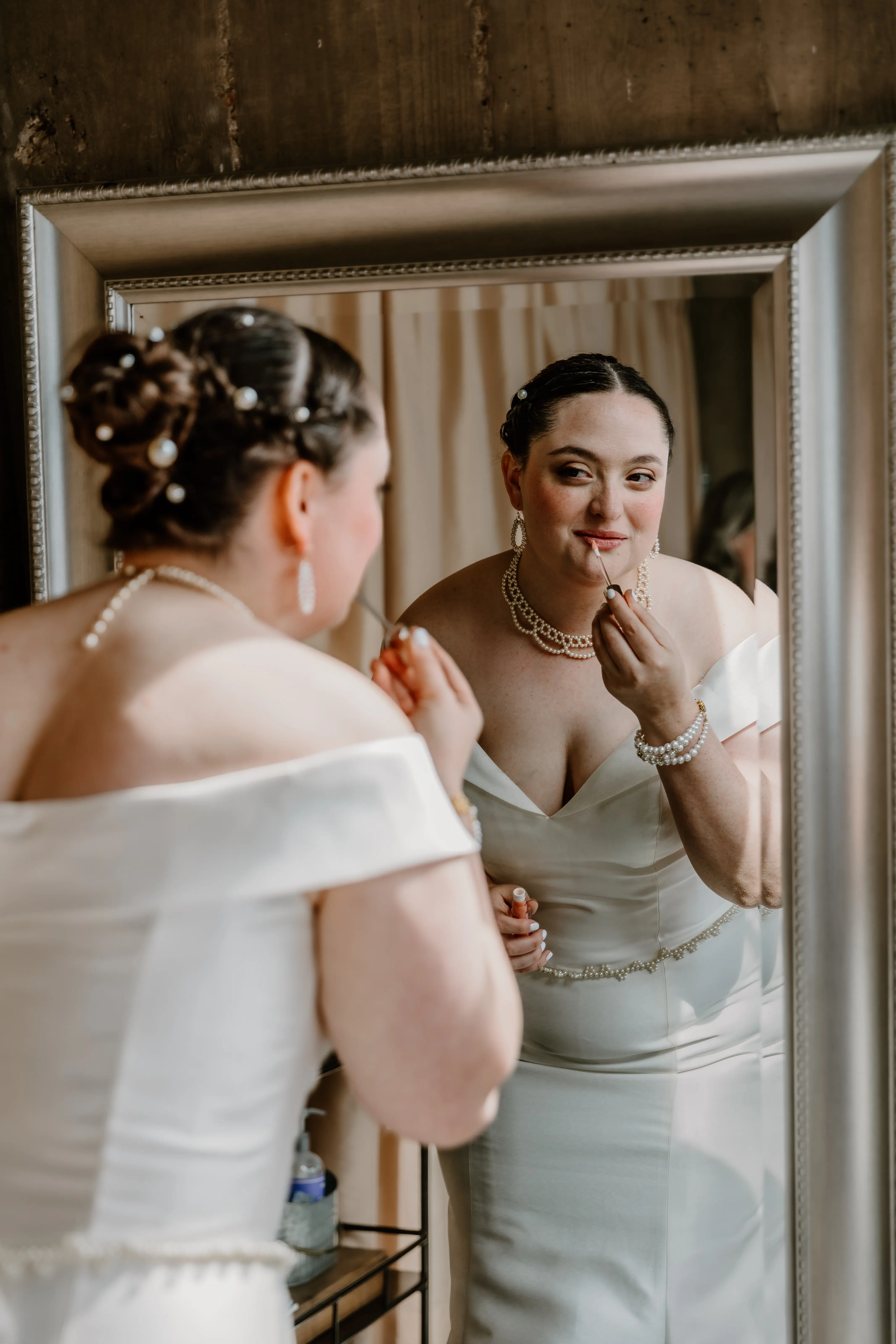 Bride Wearing Simple Fit And Flare Wedding Dress Called Josie By Rebecca Ingram Looking In A Mirror Putting On Makeup
