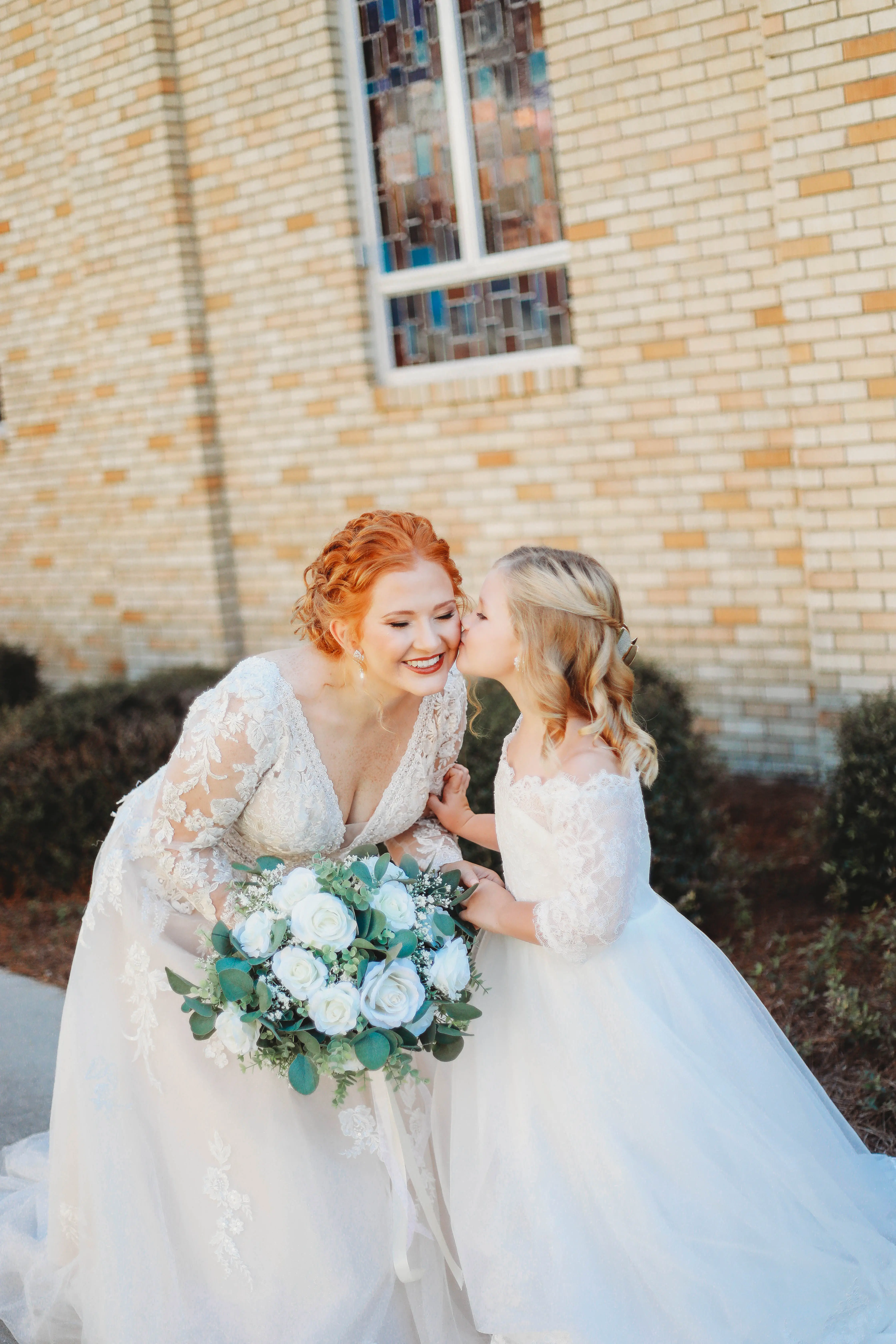 Bride Wearing Lace Long-Sleeve Wedding Gown Called Raphael By Maggie Sottero Getting Kissed By Flower Girl