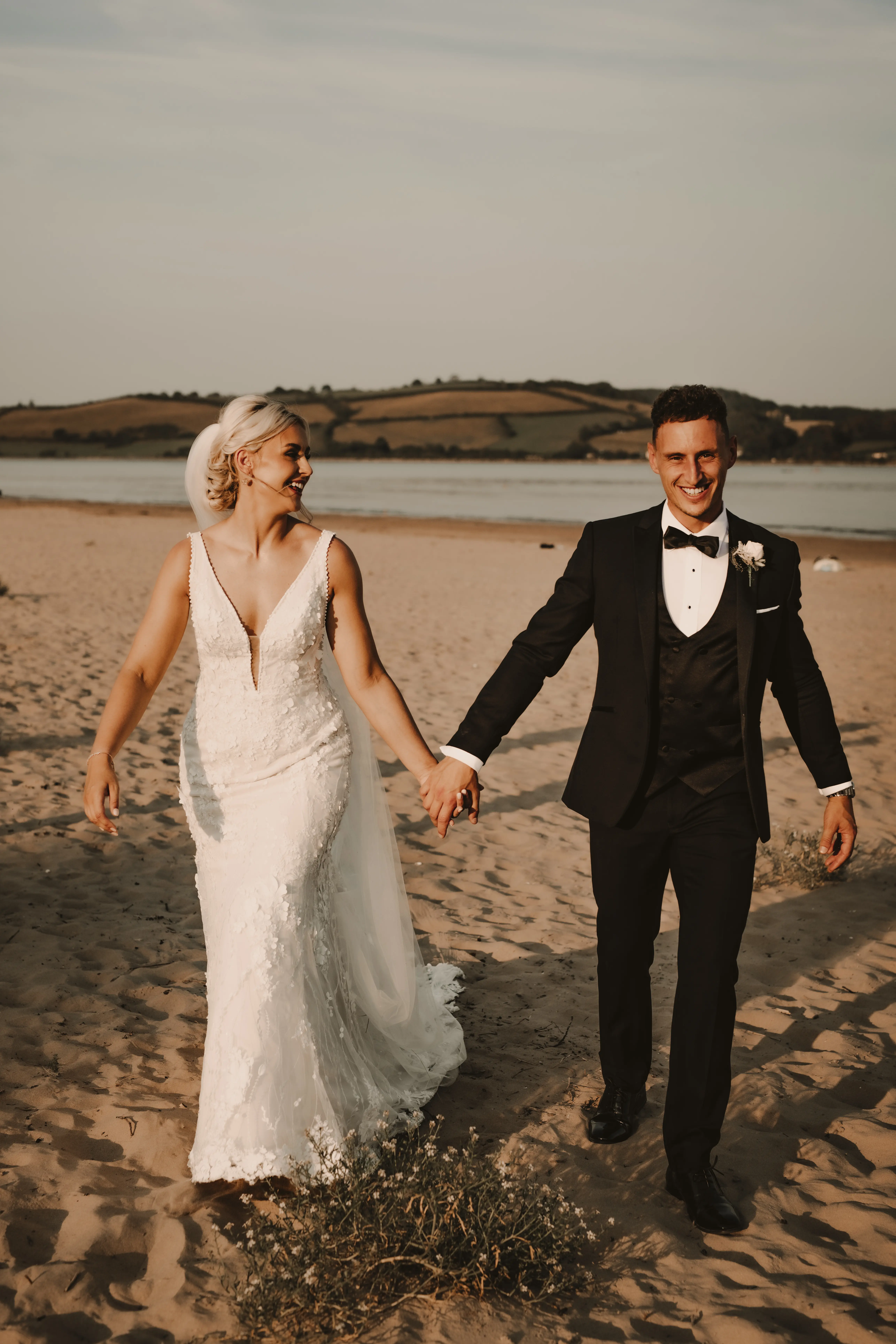 Newlyweds Holding Hands as They Walk on the Beach. Groom, Pup, and Beautiful Bride in Vintage-Inspired Mermaid Wedding Dress with 3-D Floral Motifs and Extended Scalloped Lace Train Called Cruz by Sottero & Midgley
